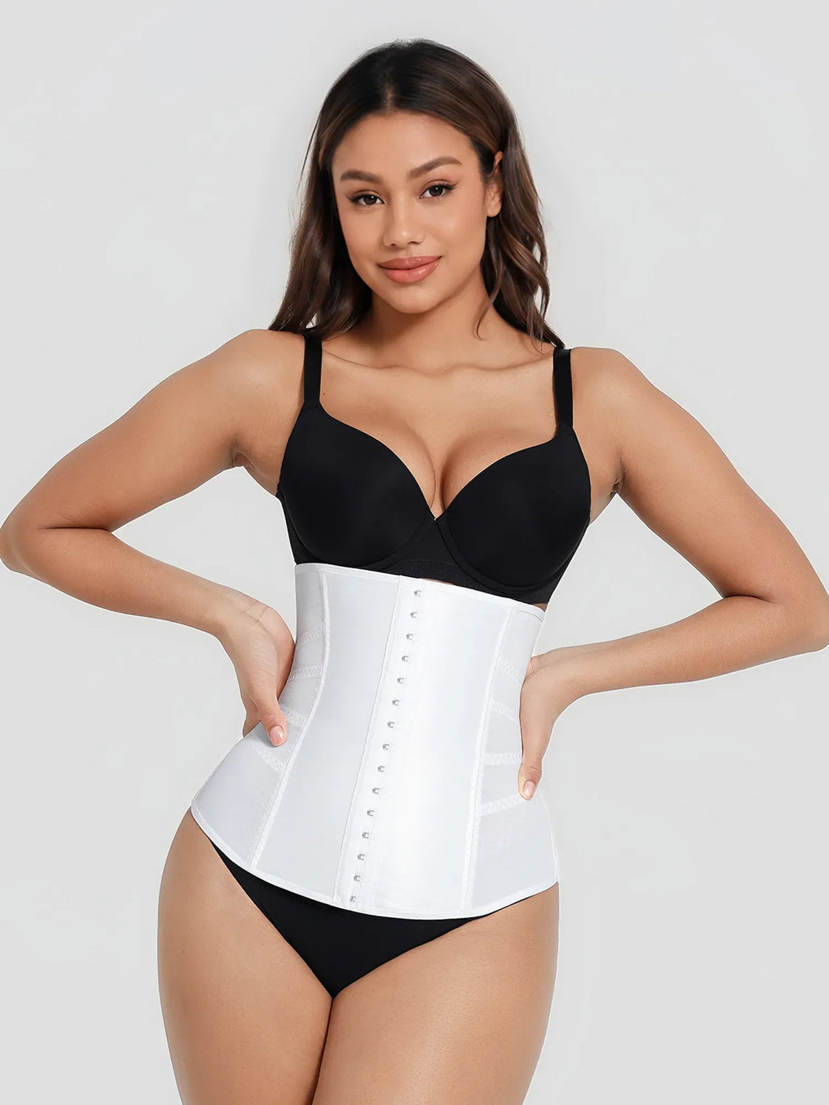 Woman wearing a black bra and white waist trainer on a plain background